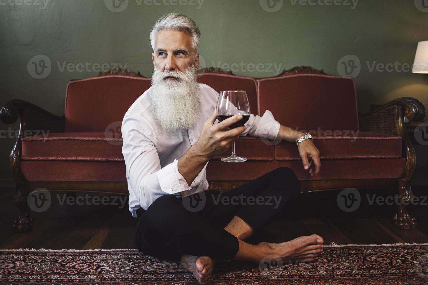 bello barbuto anziano uomo seduta su il pavimento e potabile rosso vino foto