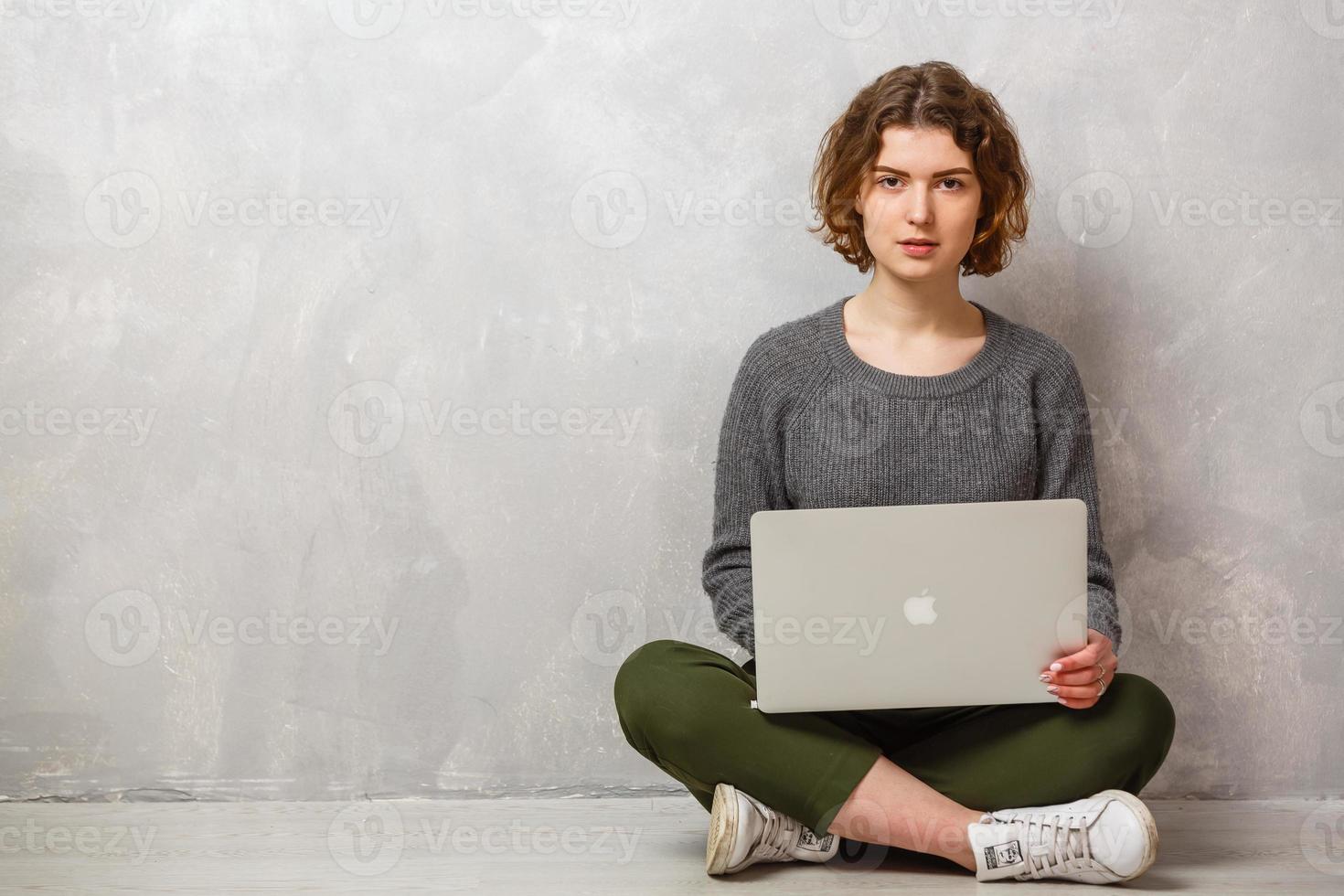 ritratto di soddisfatto femmina con bellissimo Sorridi godendo Guardando film nel argento computer e seduta nel loto posa su il pavimento al di sopra di grigio parete foto