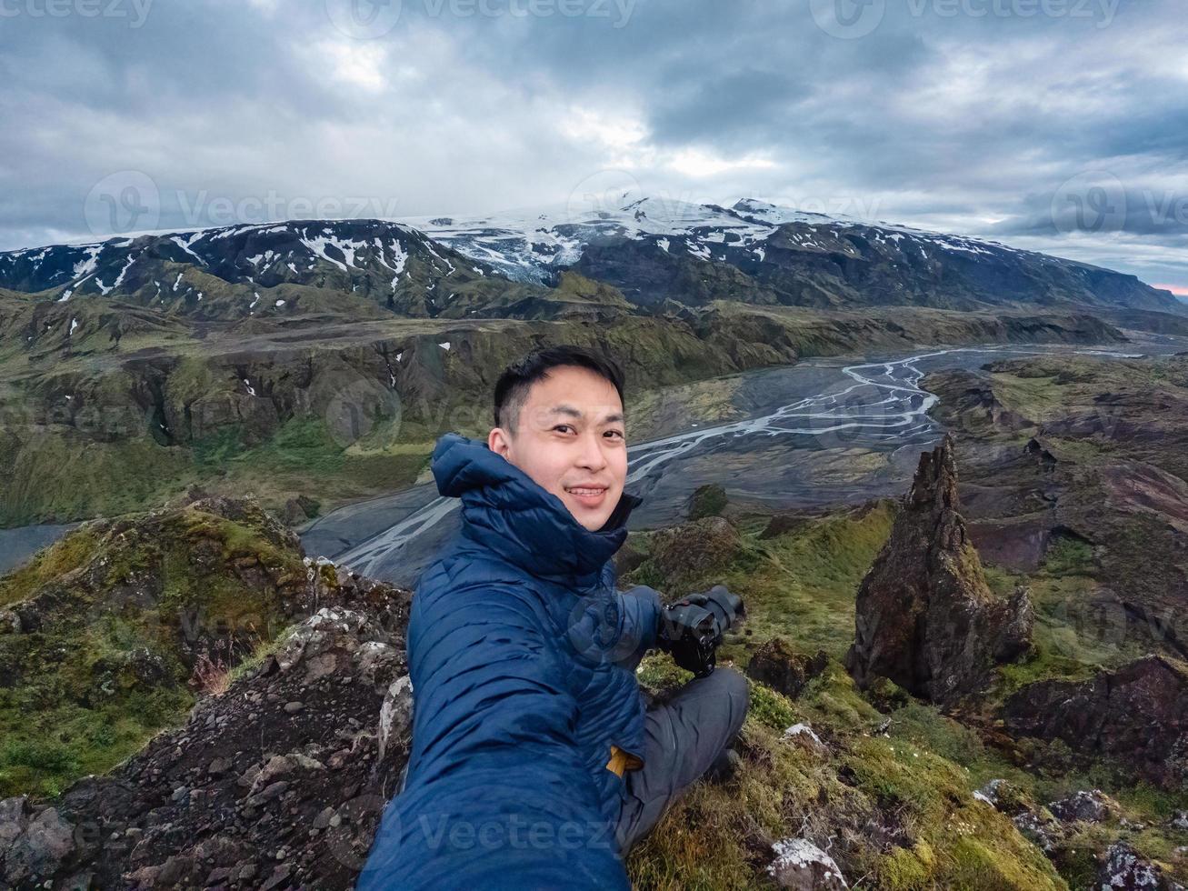 giovane asiatico maschio escursionista seduta e assunzione autoscatto su picco con montagna nel islandese Highlands su cupo giorno nel estate foto