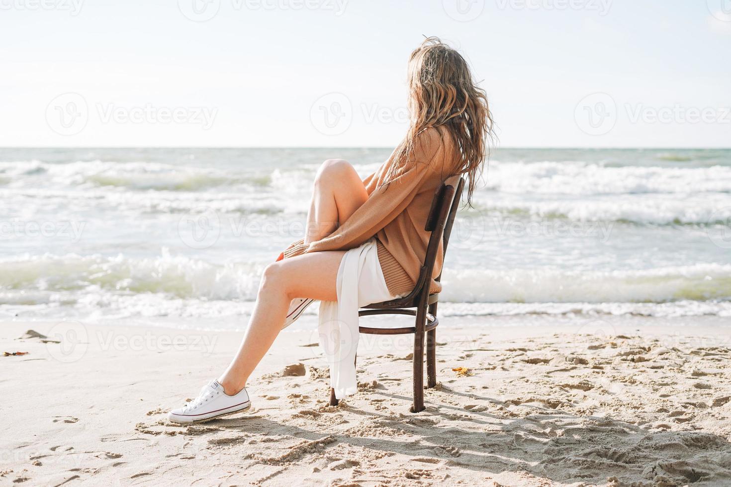 giovane spensierato bellissimo donna con lungo capelli nel maglione seduta su sedia su mare spiaggia foto