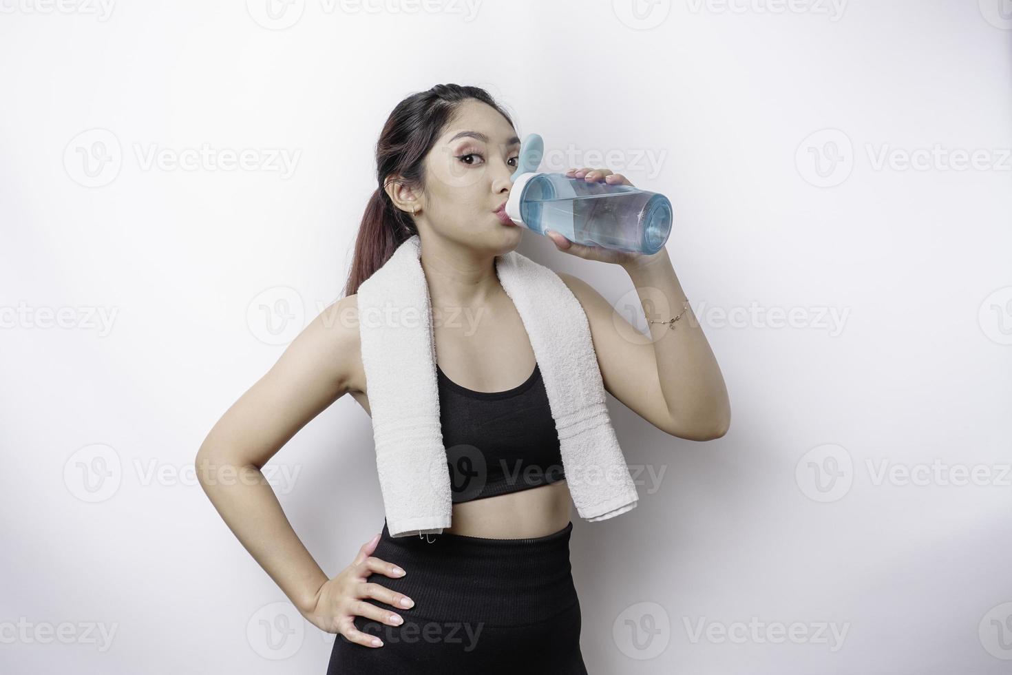 allegro asiatico donna in posa con un' asciugamano su sua spalla e potabile a partire dal un' bottiglia di acqua, sorridente e rilassante dopo allenarsi foto