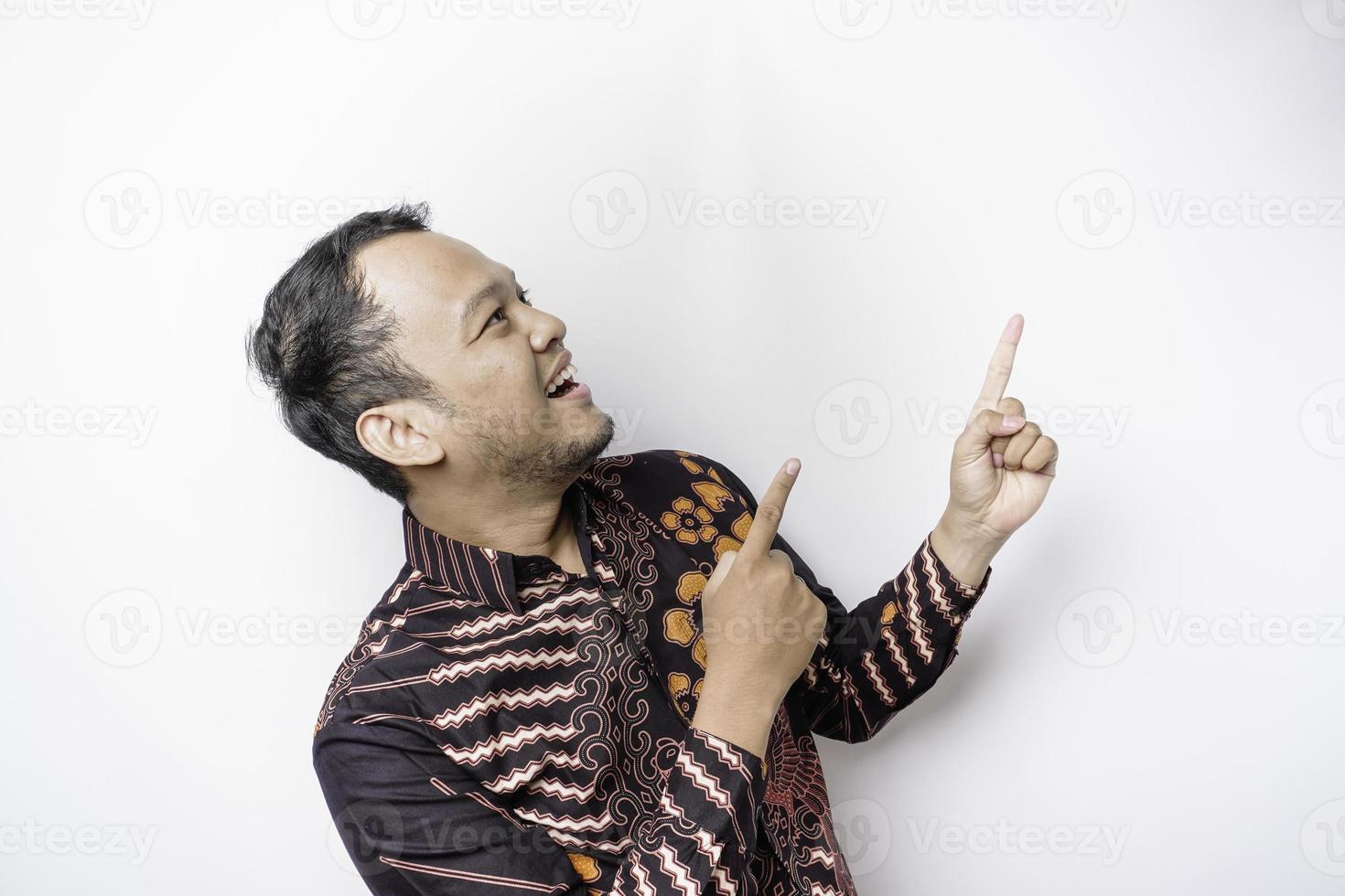 eccitato asiatico uomo indossare batik camicia puntamento a il copia spazio su superiore di lui, isolato di bianca sfondo foto