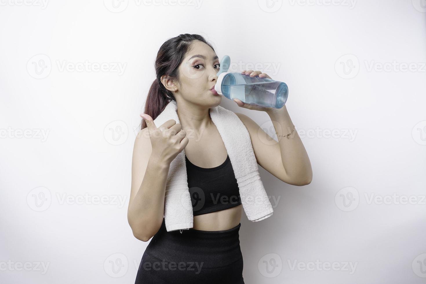 allegro asiatico donna in posa con un' asciugamano su sua spalla e potabile a partire dal un' bottiglia di acqua, sorridente e rilassante dopo allenarsi foto