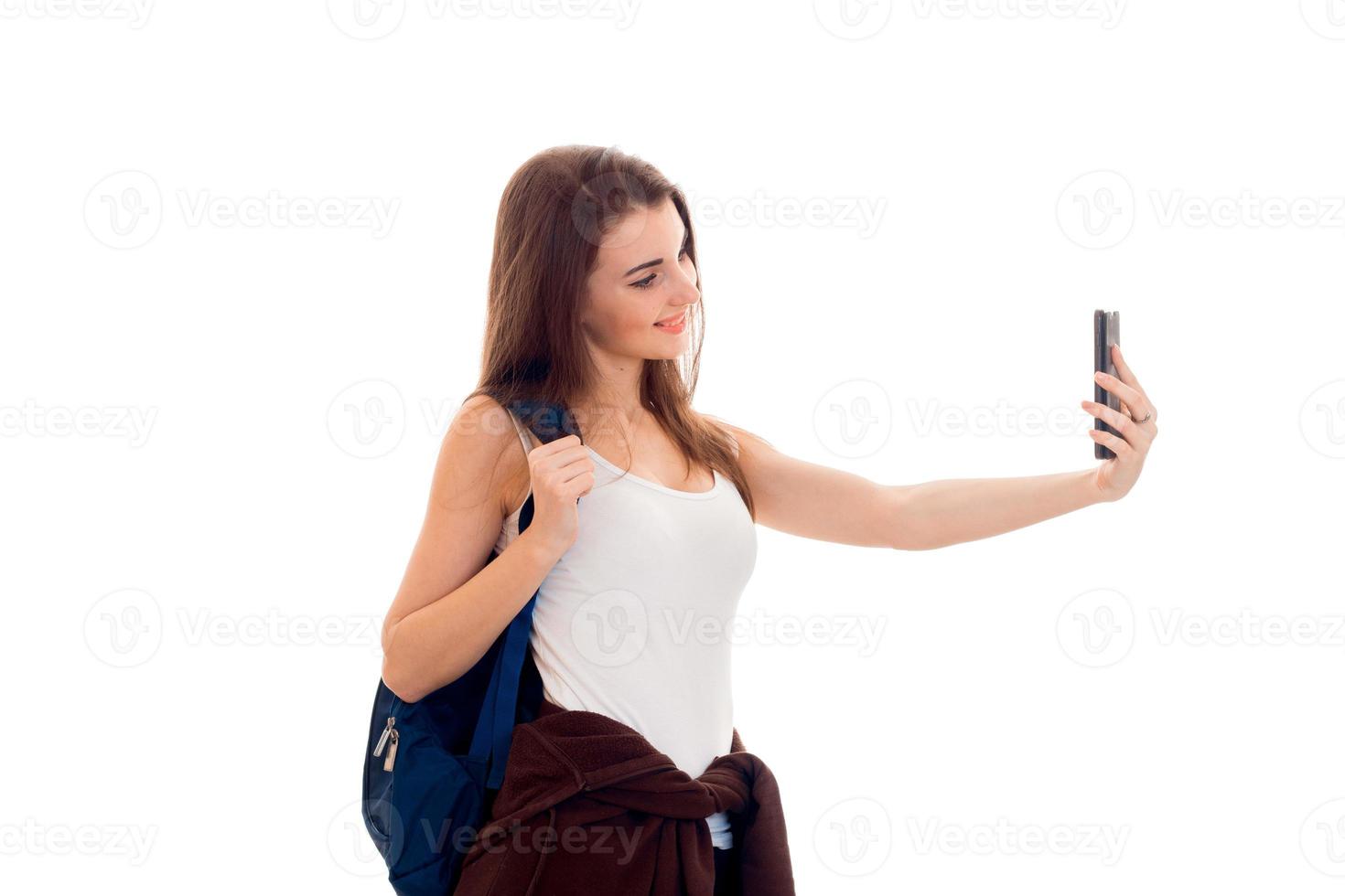 allegro giovane brunetta alunno ragazza con blu backpackand e mobile Telefono nel sua mani fa autoscatto isolato su bianca sfondo foto