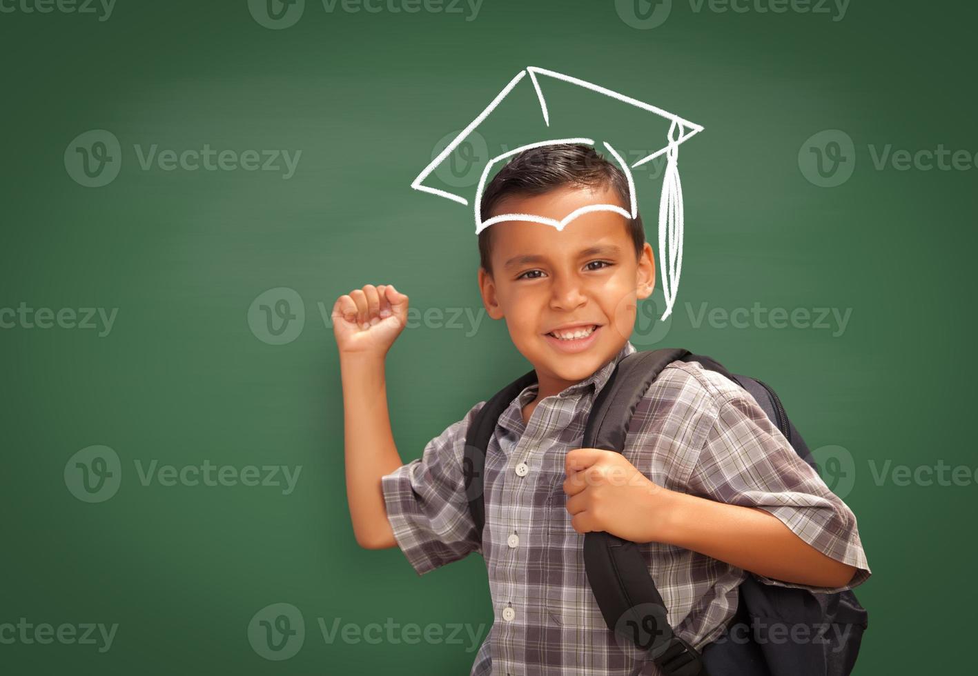 giovane ispanico alunno ragazzo indossare zaino davanti di lavagna con la laurea berretto disegnato nel gesso al di sopra di testa foto