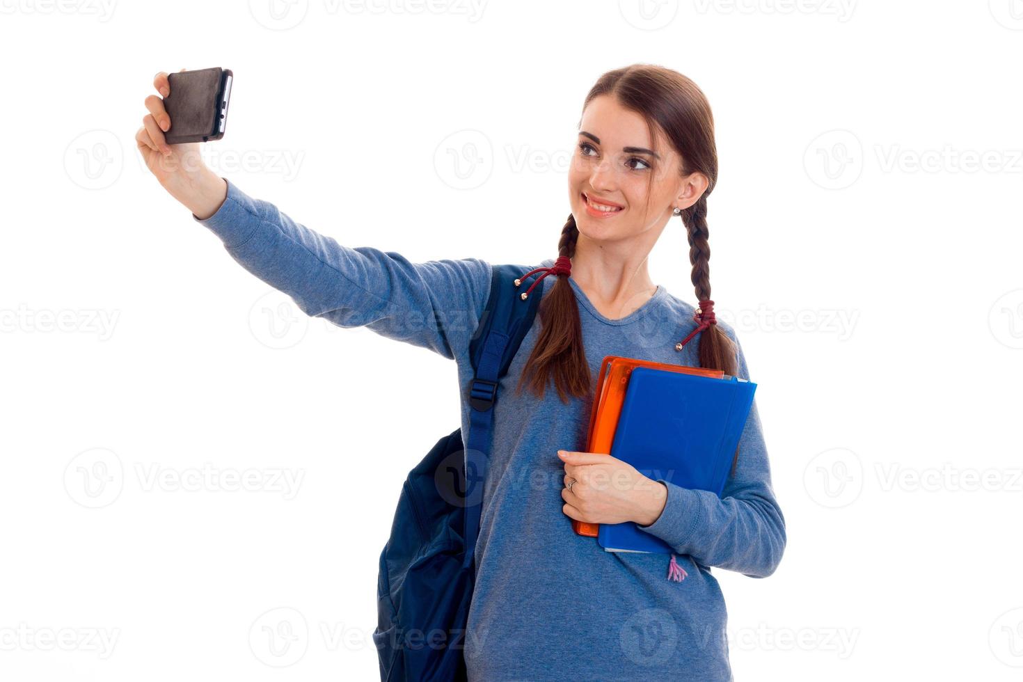 carino adolescente ragazza con trecce sollevato mano sembra in il telecamera e sorridente mentre il altro mano detiene il taccuino foto