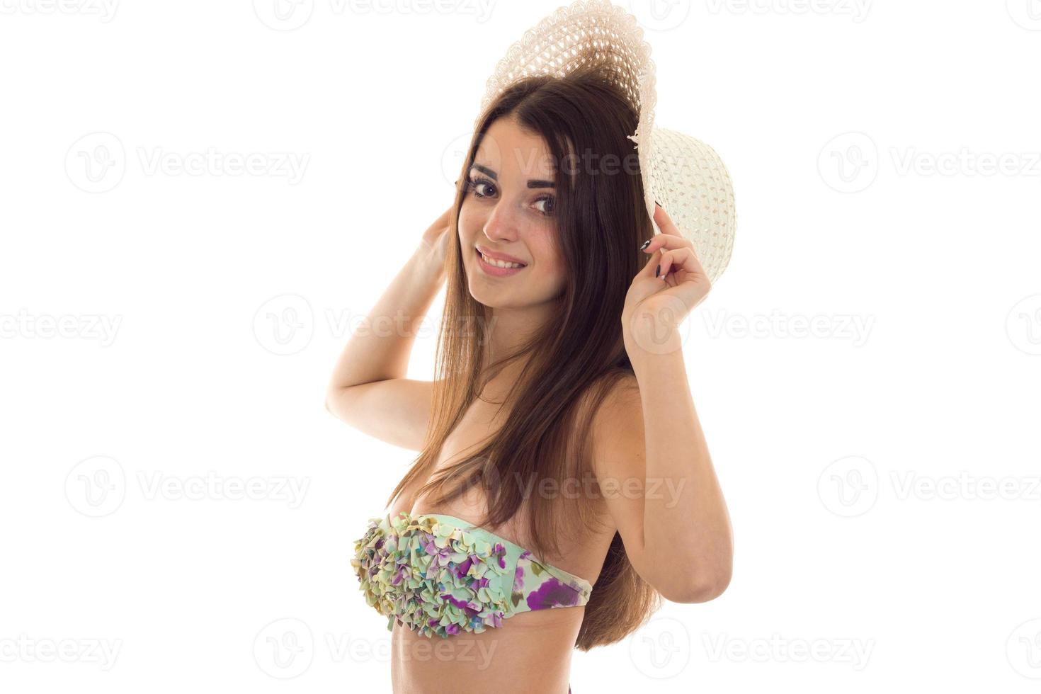 giovane carino brunetta donna con grande naturale seni nel costume da bagno con floreale modello e cannuccia cappello sorridente e guardare a il telecamera isolato su bianca sfondo foto