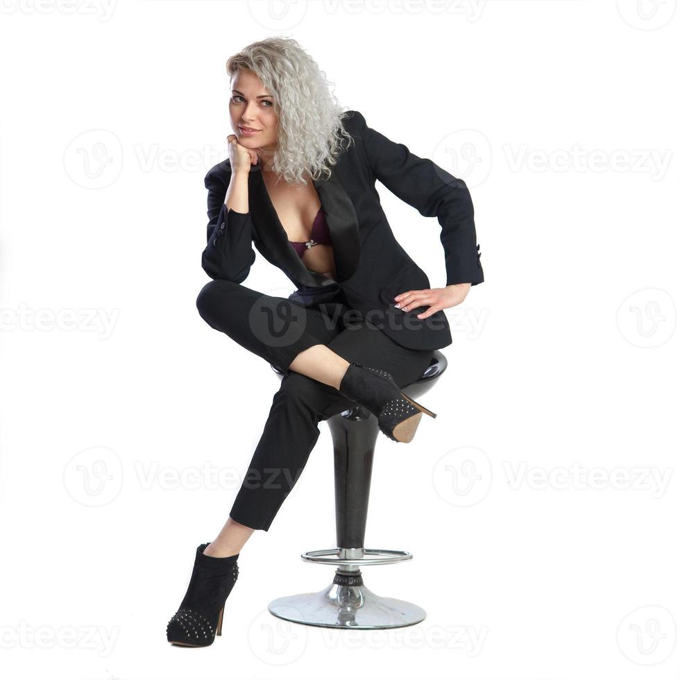 donna con bionda Riccio capelli nel nero attività commerciale completo da uomo si siede su sedia foto