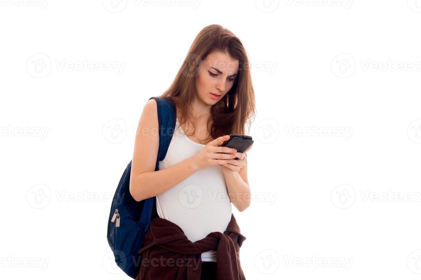 giovane bello brunetta alunno ragazza con zaino su sua le spalle taping con mobile Telefono isolato su bianca sfondo foto