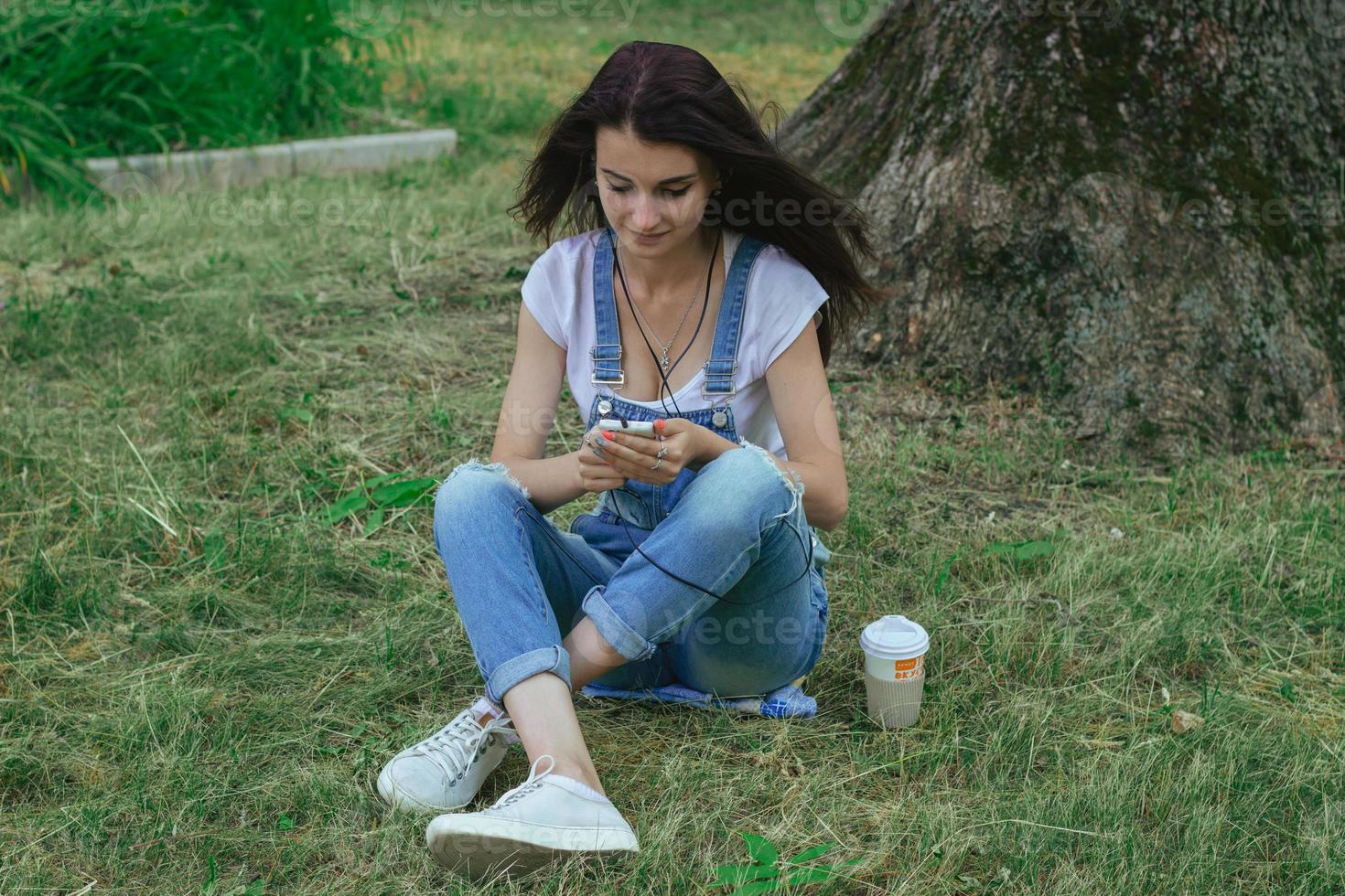 giovane affascinante brunetta si siede nel il erba ascoltando per musica e guardare a Telefono foto