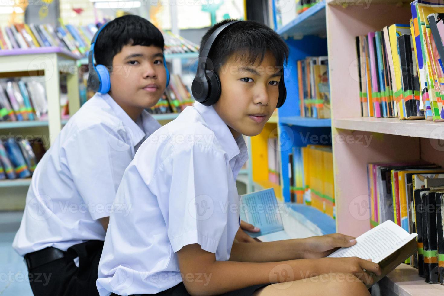 morbido fucus di Due asiatico ragazzo studenti siamo ascoltando media, lettura e consulenza di preferito libro a biblioteca di scuola foto