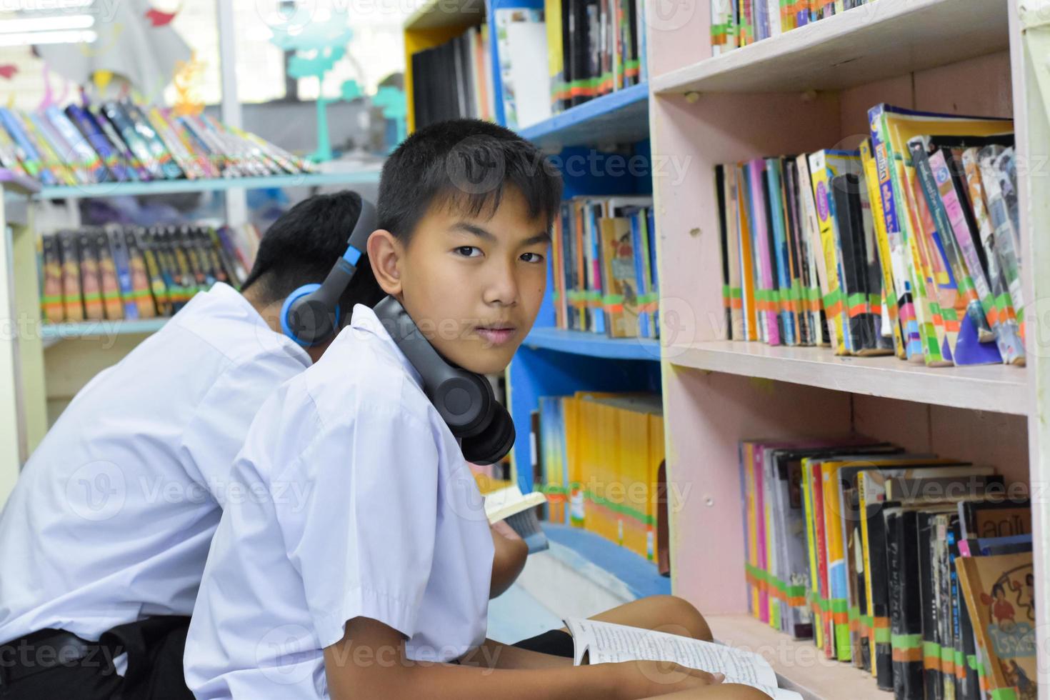 morbido fucus di Due asiatico ragazzo studenti siamo ascoltando media, lettura e consulenza di preferito libro a biblioteca di scuola foto