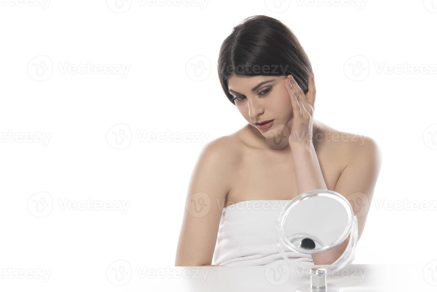 bellissimo attraente brunetta donna controllo sua viso pelle nel davanti di un' specchio su bianca sfondo foto