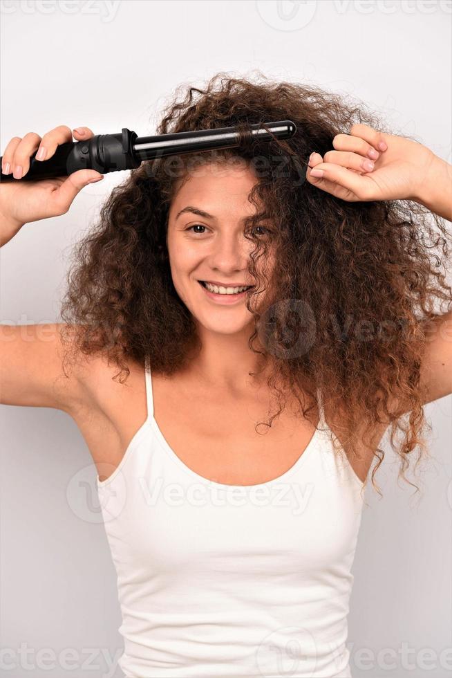 bellissimo sorridente donna con lungo ondulato capelli Stirare esso, utilizzando arricciatura ferro da stiro. contento ragazza con bellissima salutare liscio capelli utilizzando bigodino per Perfetto riccioli. acconciatura e parrucchiere foto