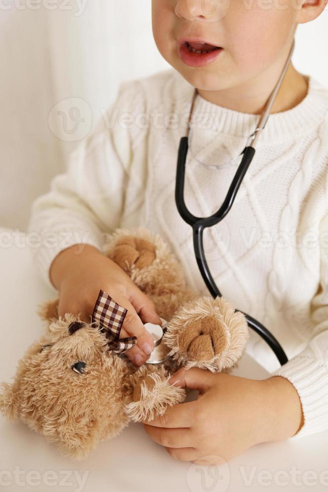 carino poco ragazzo giocando medico a casa e curare felpa giocattolo. dolce bambino piccolo bambino utilizzando stetoscopio. avendo divertimento. bambini e medicinale, assistenza sanitaria. foto