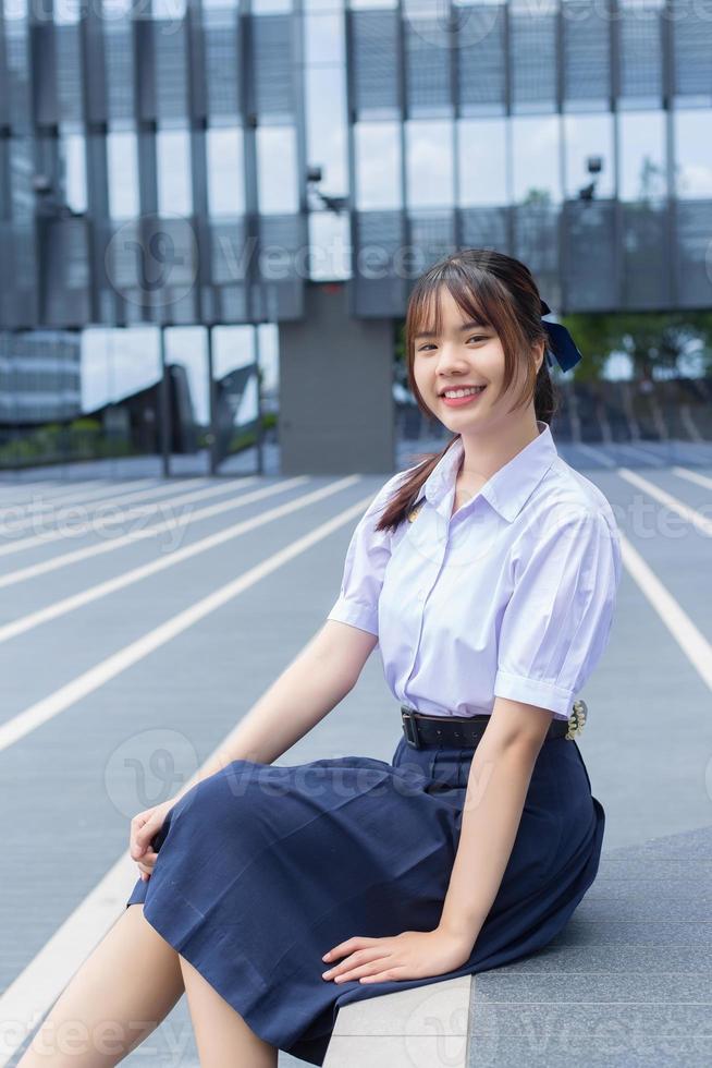 carino asiatico alto scuola alunno ragazza nel il scuola uniforme con sorrisi con fiducia mentre lei sembra a il telecamera felicemente con il edificio nel il sfondo. foto