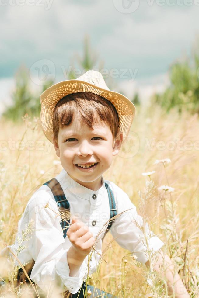 poco contadino su un' estate campo, carino poco ragazzo nel un' cannuccia cappello. ragazzo nel un' cappello con un' fiore sta nel un' campo. ritratto di un' villaggio ragazzo foto