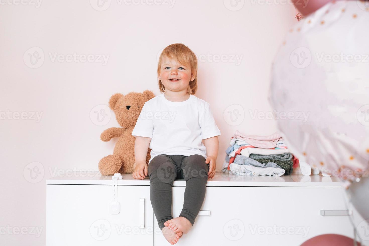 carino divertente bambino ragazza contento bambino nel casa Abiti seduta su cassettone a casa foto