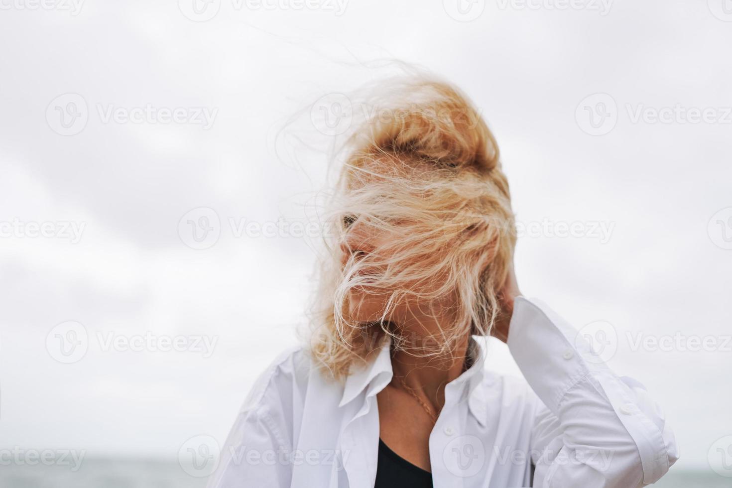 ritratto di elegante bionda donna nel bianca camicia su sabbia spiaggia a tempesta mare a ventoso weater foto