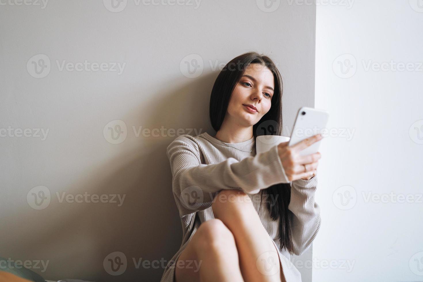 bellissimo sorridente donna adolescente ragazza alunno con buio lungo capelli nel casa Abiti con mattina tazza di caffè o tè utilizzando mobile Telefono accogliente media su il grigio sfondo foto