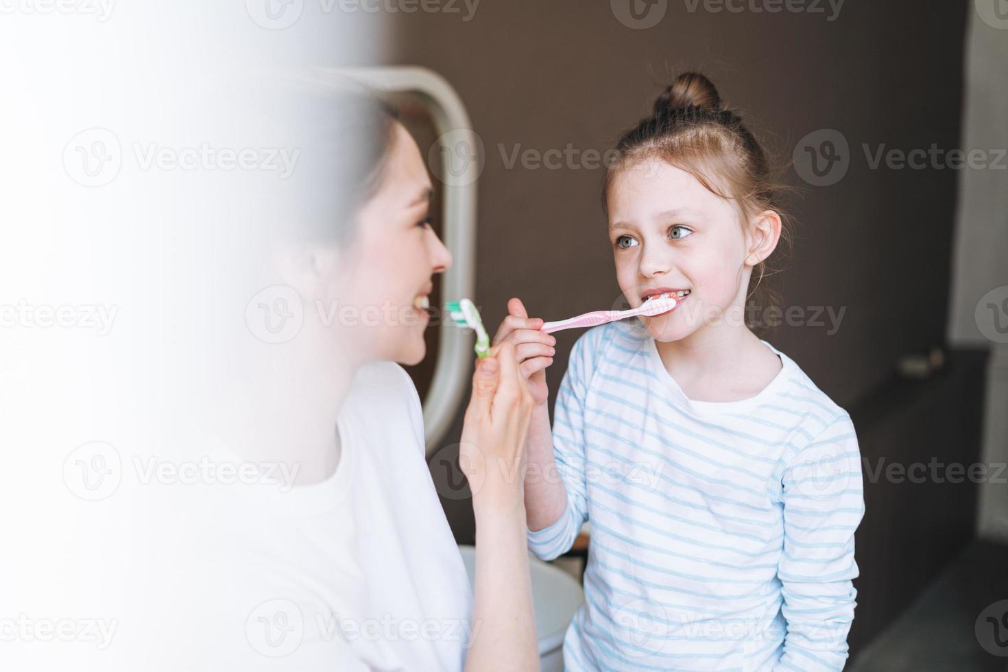 giovane madre donna con lungo capelli con poco gemello ragazza figlia nel pigiama spazzolatura loro denti nel il mattina a casa foto