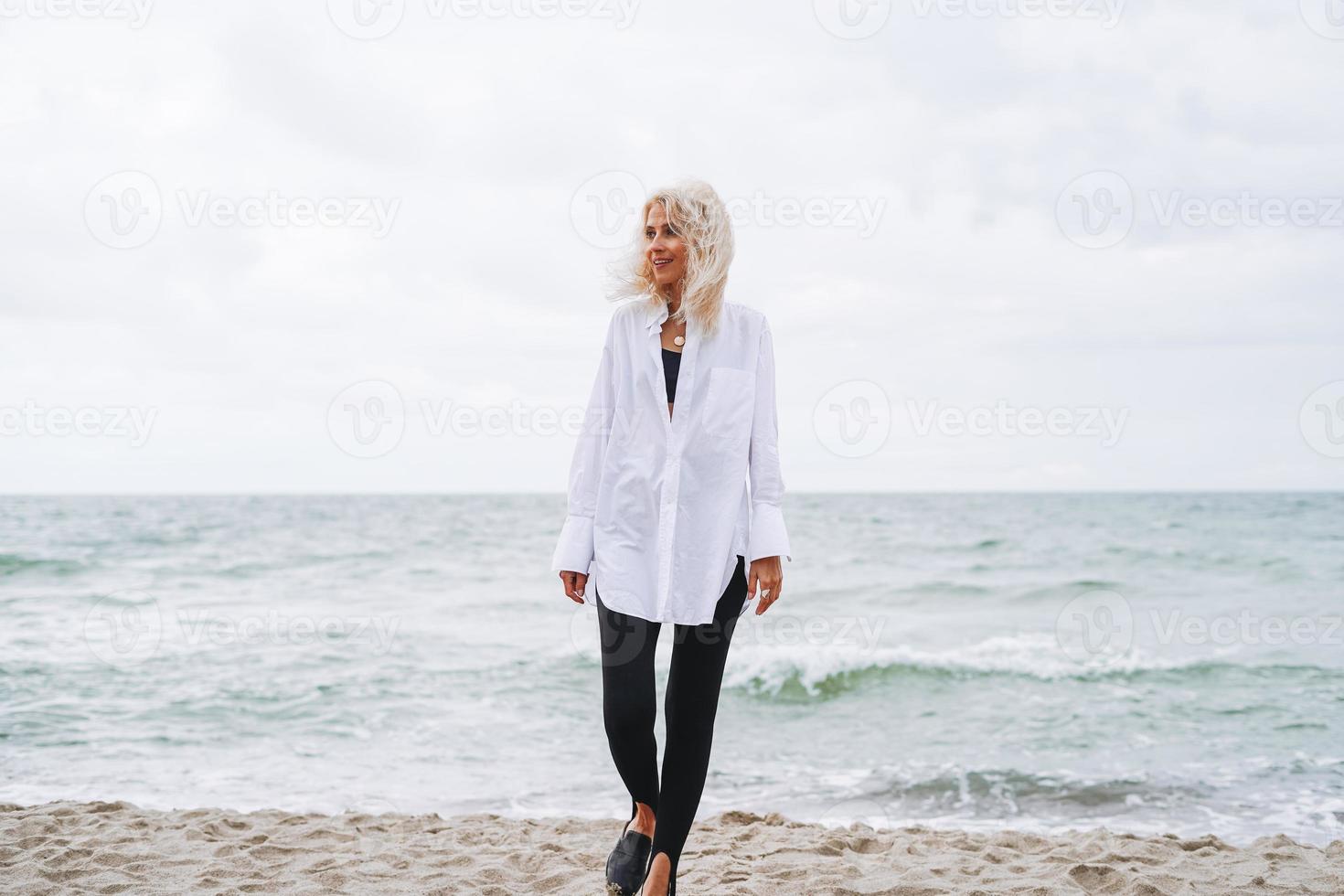 ritratto di elegante bionda donna nel bianca camicia su sabbia spiaggia a tempesta mare a ventoso weater foto