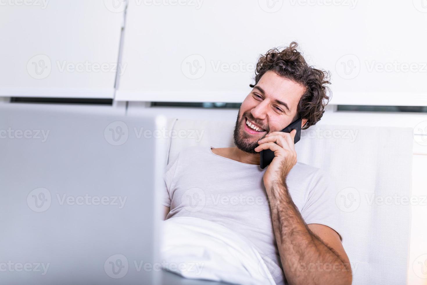 Immagine di giovane caucasico uomo ha mattina caffè, si siede su letto, Lavorando su il suo il computer portatile e parlando su il suo mobile Telefono, prepara per visita medica, usi il computer portatile computer per fare surf Internet. foto