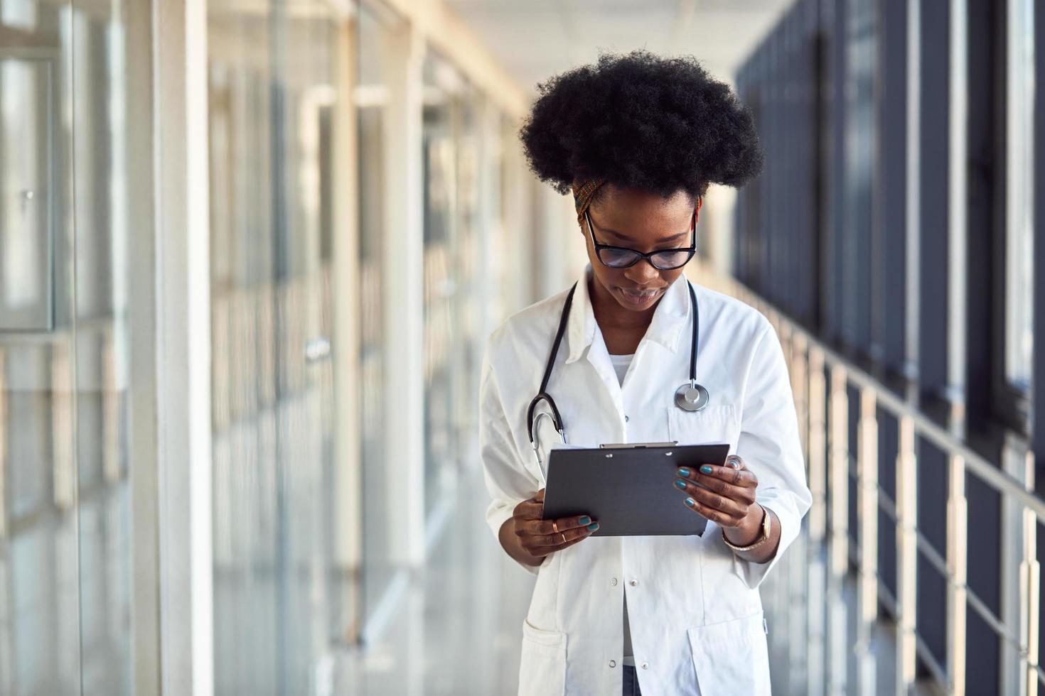 giovane africano americano femmina medico nel bianca uniforme con stetoscopio e bloc notes in piedi nel corridoio foto