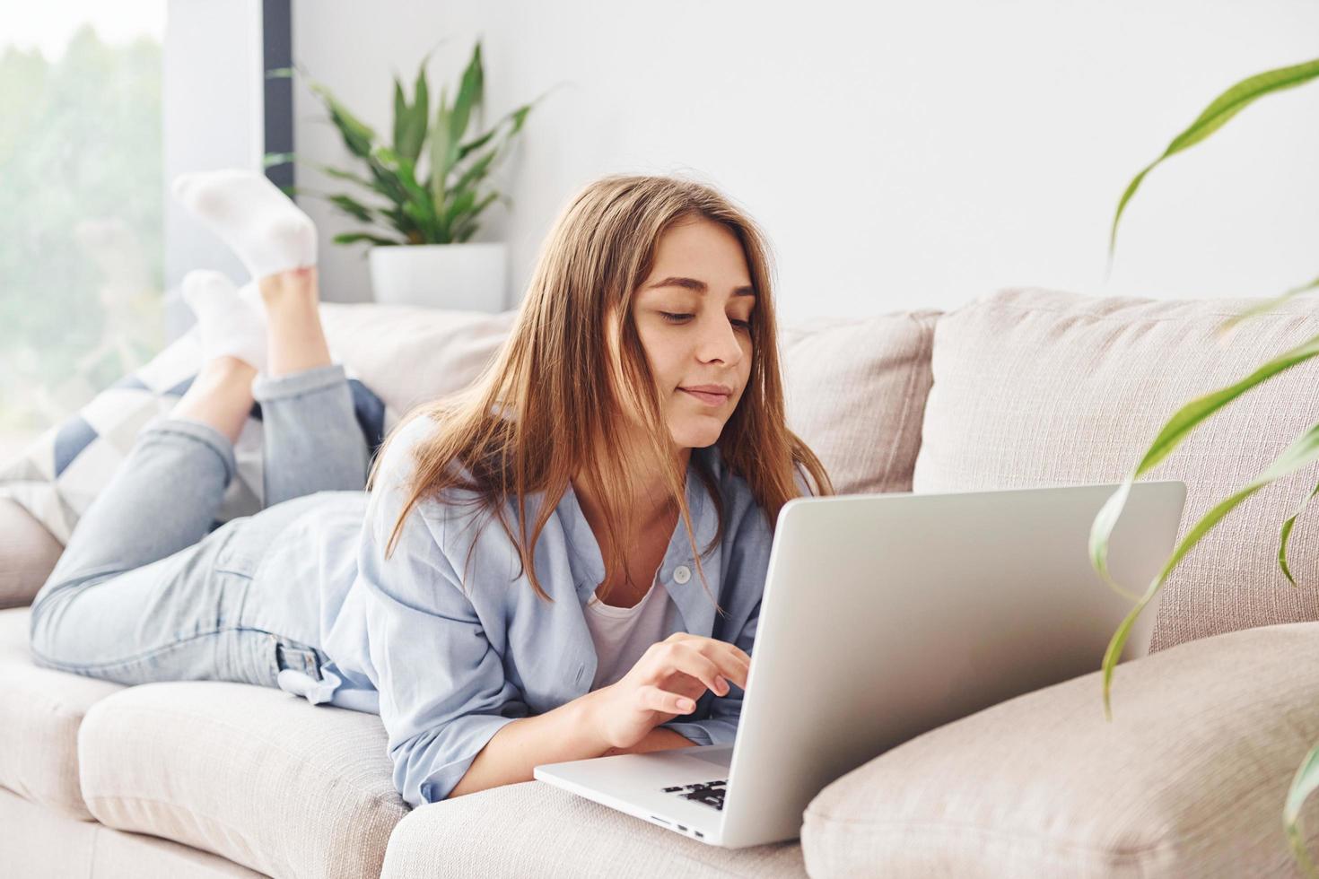 bellissimo giovane donna nel jeans e blu camicia dire bugie giù a divano con il computer portatile foto