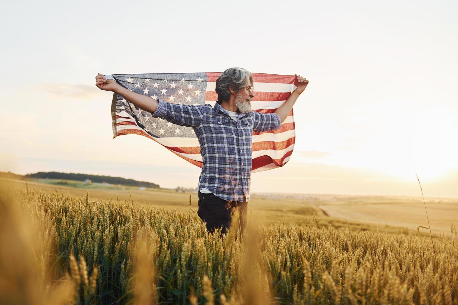 Tenere Stati Uniti d'America bandiera nel mani. patriottico anziano elegante uomo con grigio capelli e barba su il agricolo campo foto