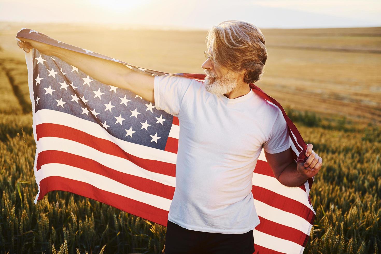 concezione di libertà. Tenere Stati Uniti d'America bandiera. anziano elegante uomo con grigio capelli e barba su il agricolo campo con raccogliere foto