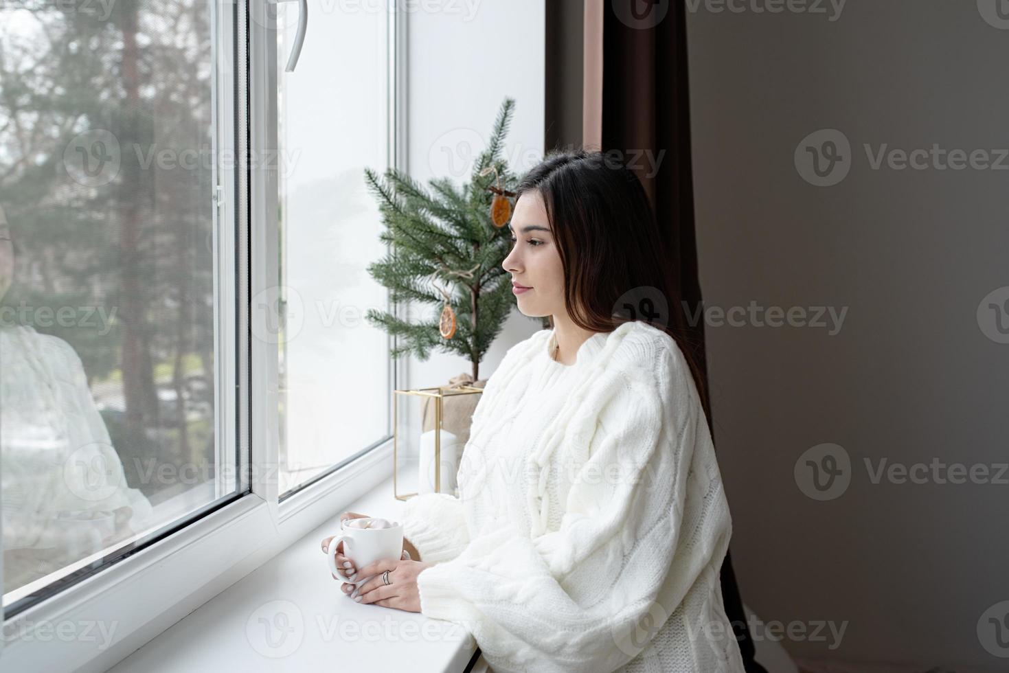 donna nel caldo bianca inverno maglione in piedi Il prossimo per il finestra a casa a Natale vigilia Tenere tazza con marshmallows, abete albero dietro a foto