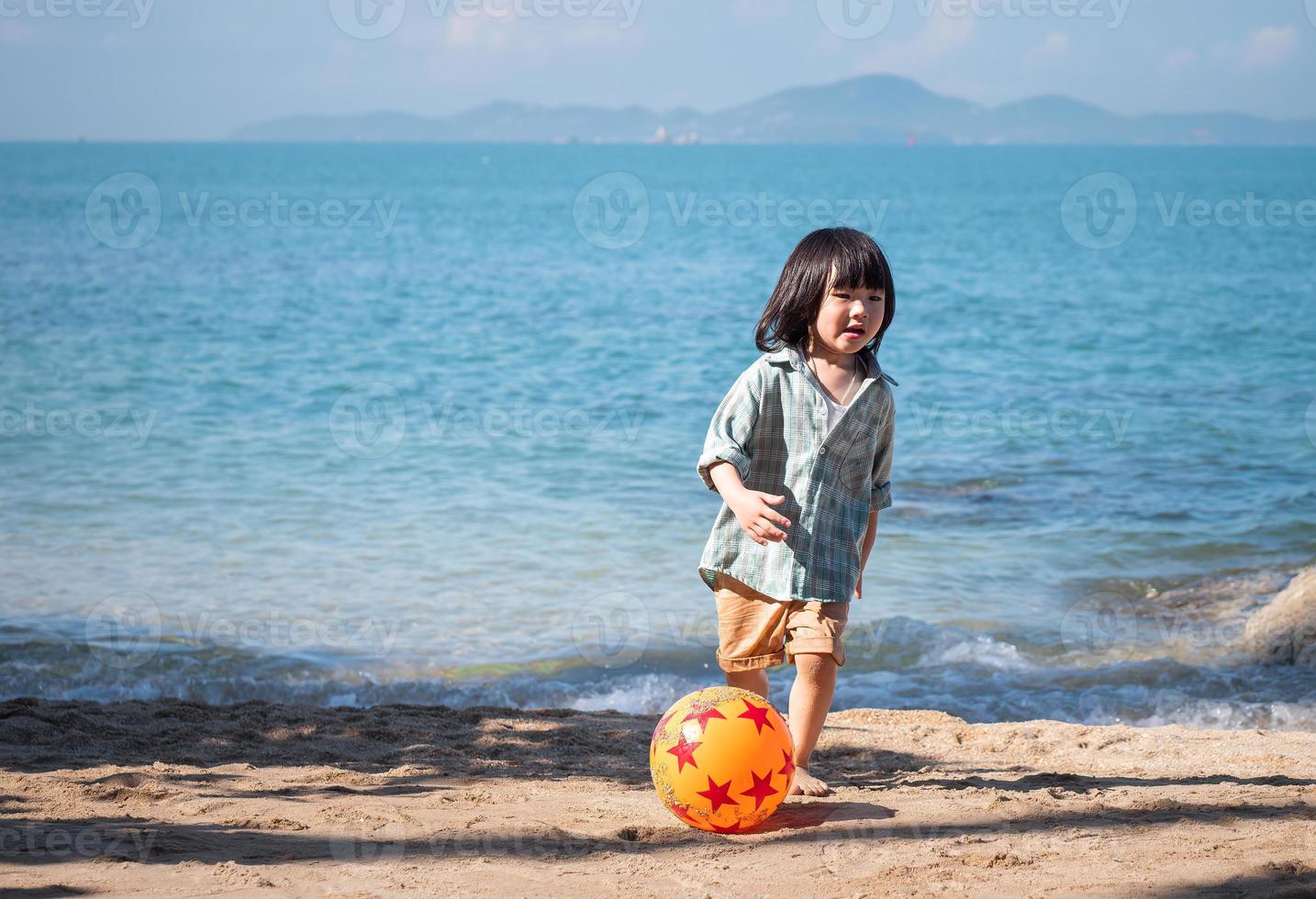 contento asiatico poco ragazzo giocando a piedi nudi calcio su il spiaggia con un' rosso e arancia palla nel il giorno, ragazzo avendo divertimento all'aperto. concetto di estate vacanza. foto