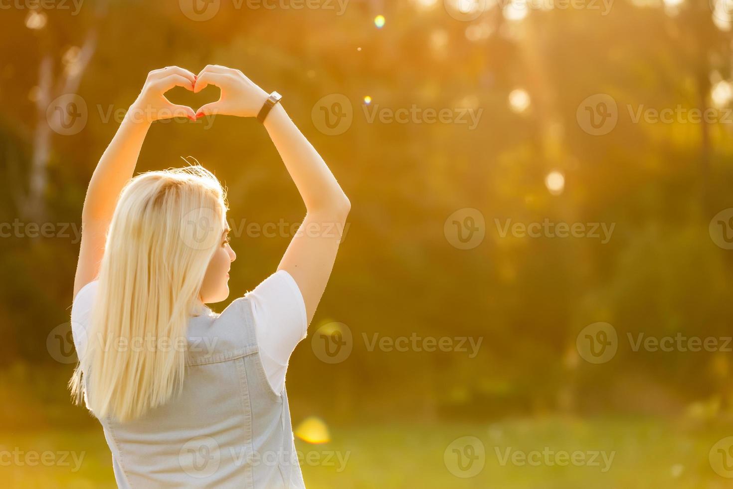 un' giovane ragazza fabbricazione cuore simbolo con sua mani a tramonto foto