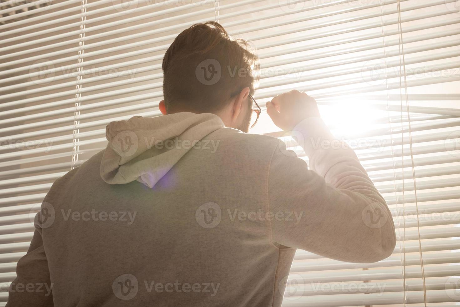 posteriore Visualizza di giovane elegante uomo sbirciando attraverso buco nel finestra persiane e guardare su in strada. concetto di godendo il mattina sole e positività foto