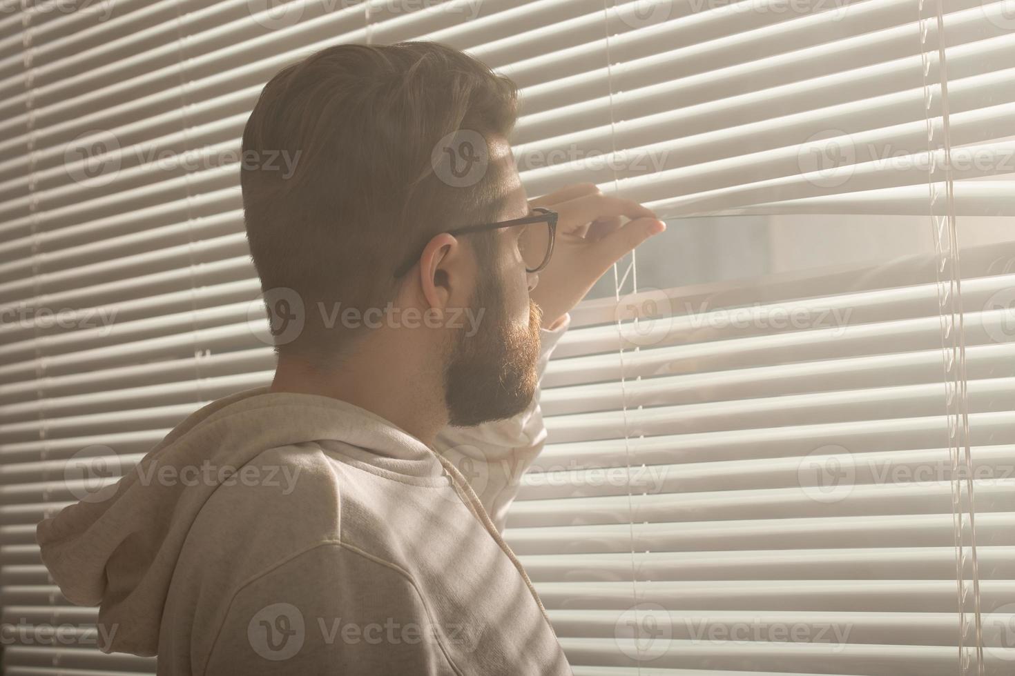 posteriore Visualizza di giovane uomo con barba sbircia attraverso buco nel il finestra persiane e sembra su in il strada. sorveglianza e curiosità concetto foto