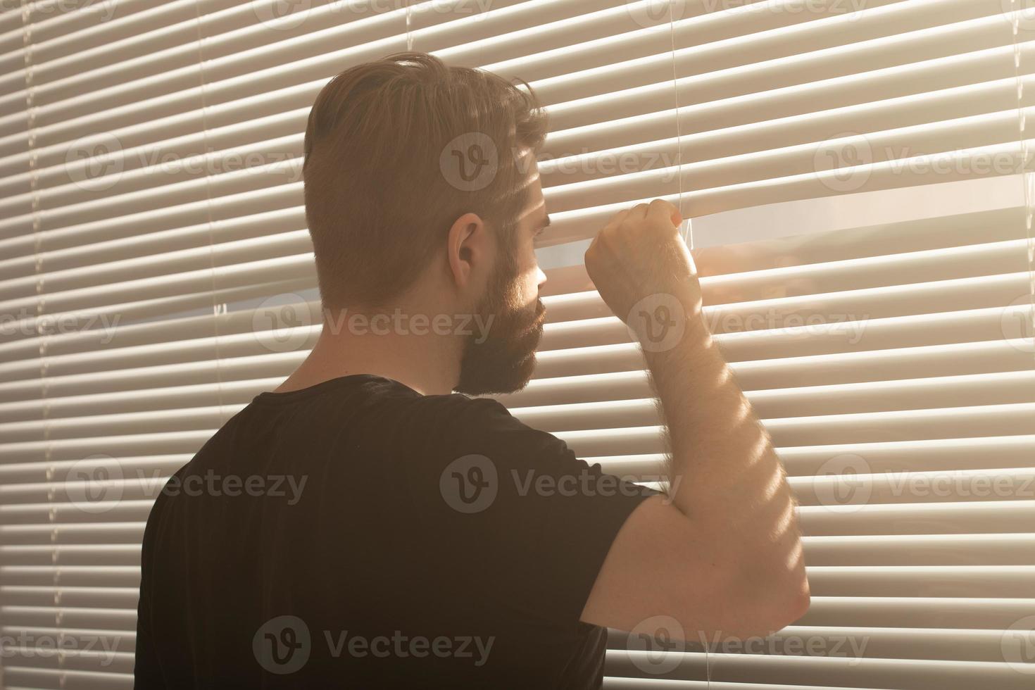 posteriore Visualizza di giovane uomo con barba sbircia attraverso buco nel il finestra persiane e sembra su in il strada. sorveglianza e curiosità concetto foto