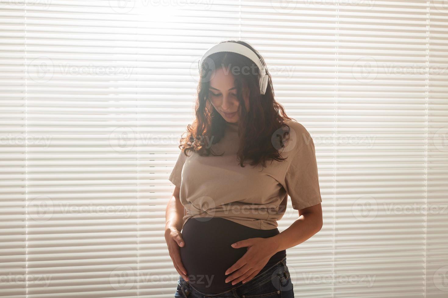 sorridente brunetta pacificato incinta donna ascoltando per piacevole musica utilizzando un' smartphone e cuffia. sedazione terapia concetto prima incontro con bambino foto