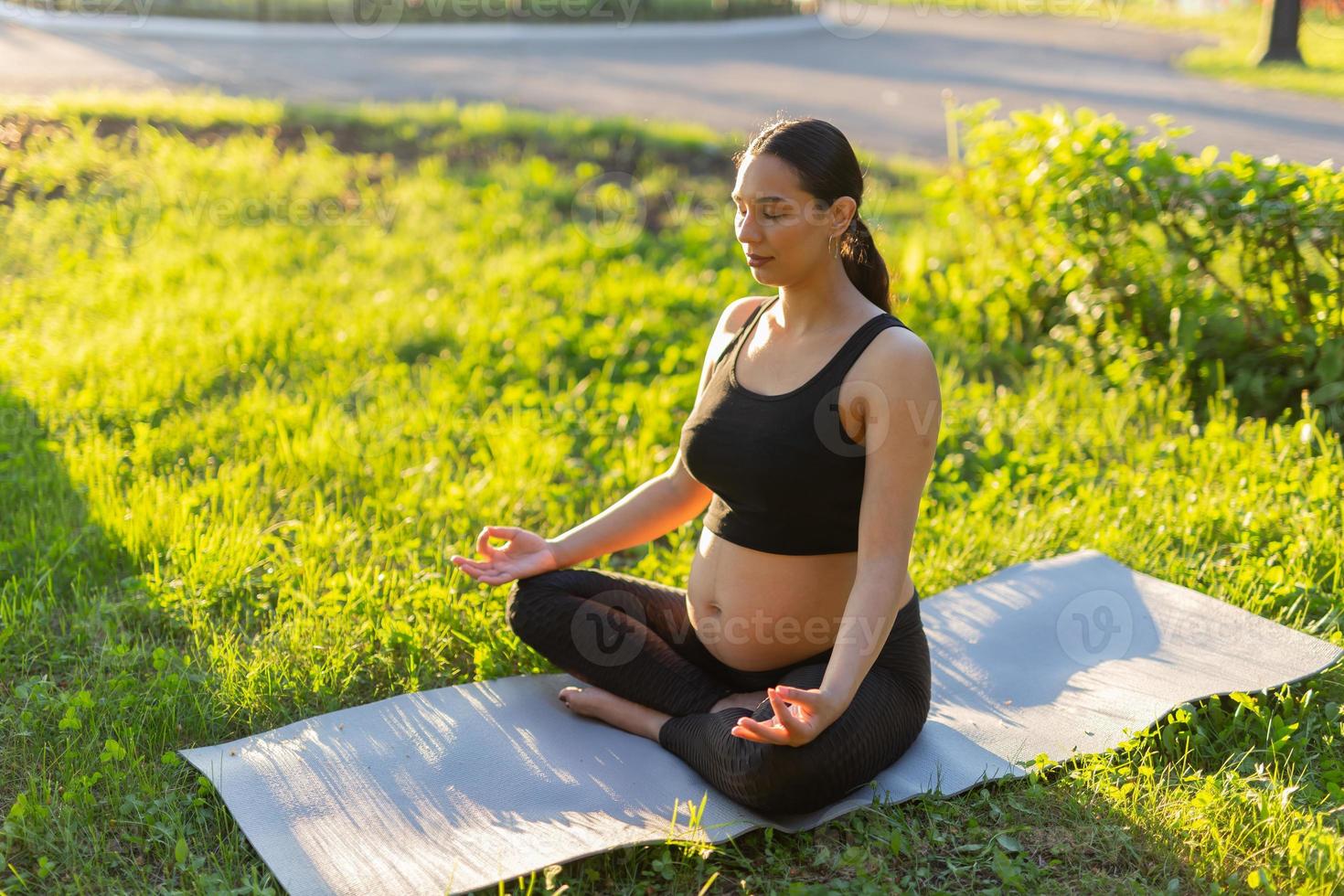 tranquillo, calmo giovane positivo incinta donna nel ginnastica completo da uomo fa yoga e meditare seduta su stuoia su verde erba su soleggiato caldo estate giorno. concetto di preparazione per parto e positivo atteggiamento foto