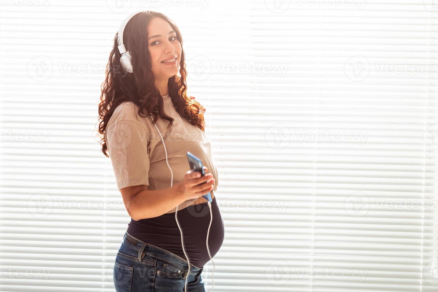 dai capelli ricci brunetta pacificato incinta donna ascolta per piacevole classico musica utilizzando smartphone e cuffia. concetto di un' calmante umore prima incontro bambino. copyspace foto