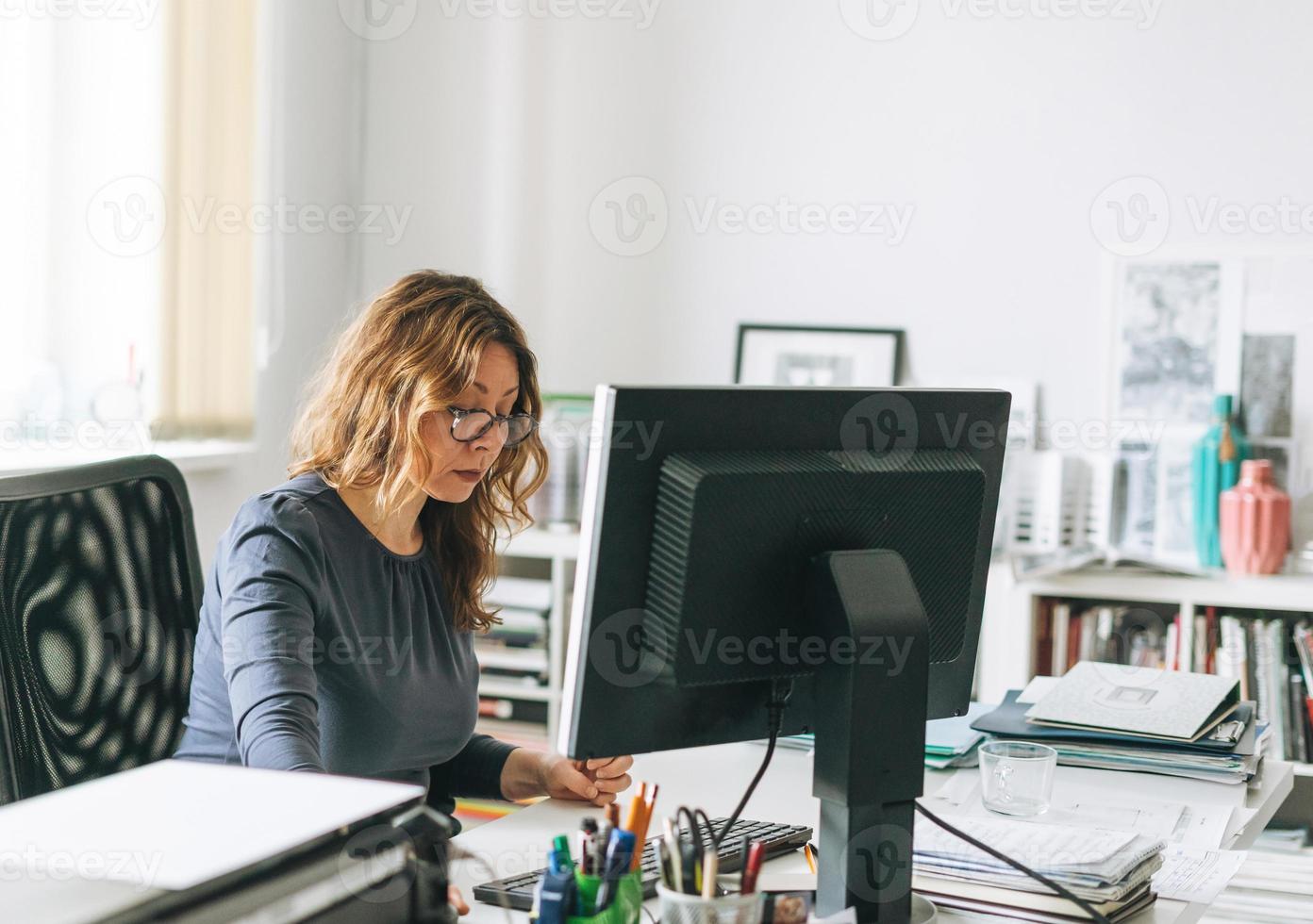 affascinante sorridente adulto donna mezzo anziano donna con Riccio capelli architetto progettista Lavorando su computer nel il luminosa moderno ufficio foto