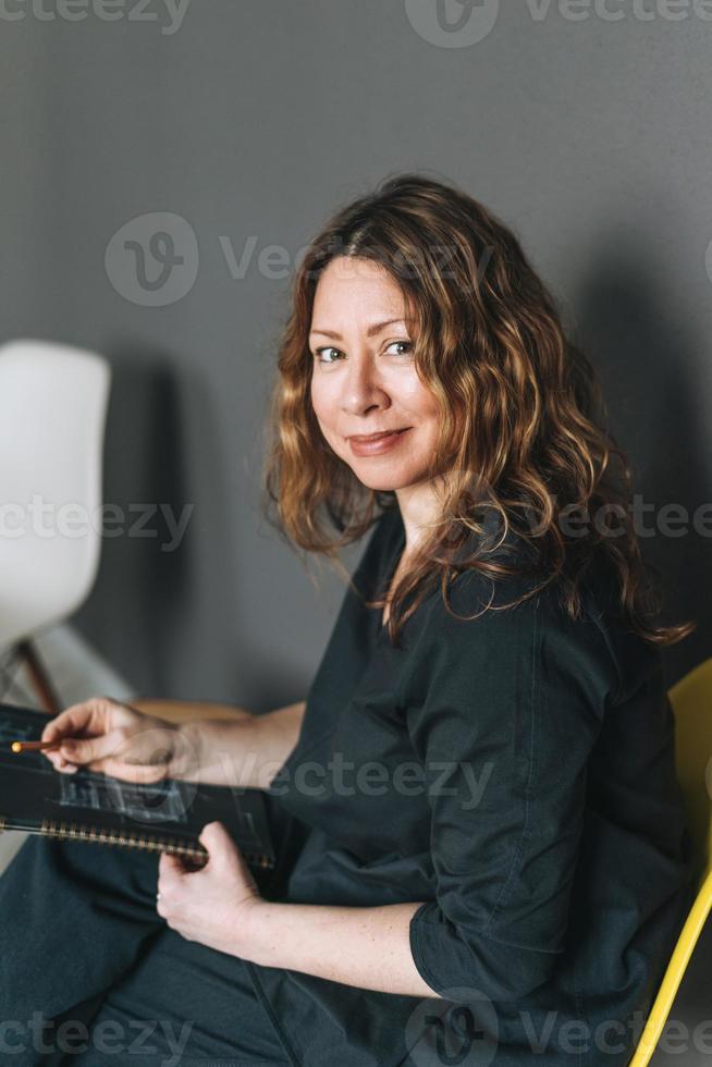 affascinante sorridente adulto donna mezzo anziano donna con Riccio capelli architetto progettista con disegno Piano nel mani nel il moderno ufficio foto