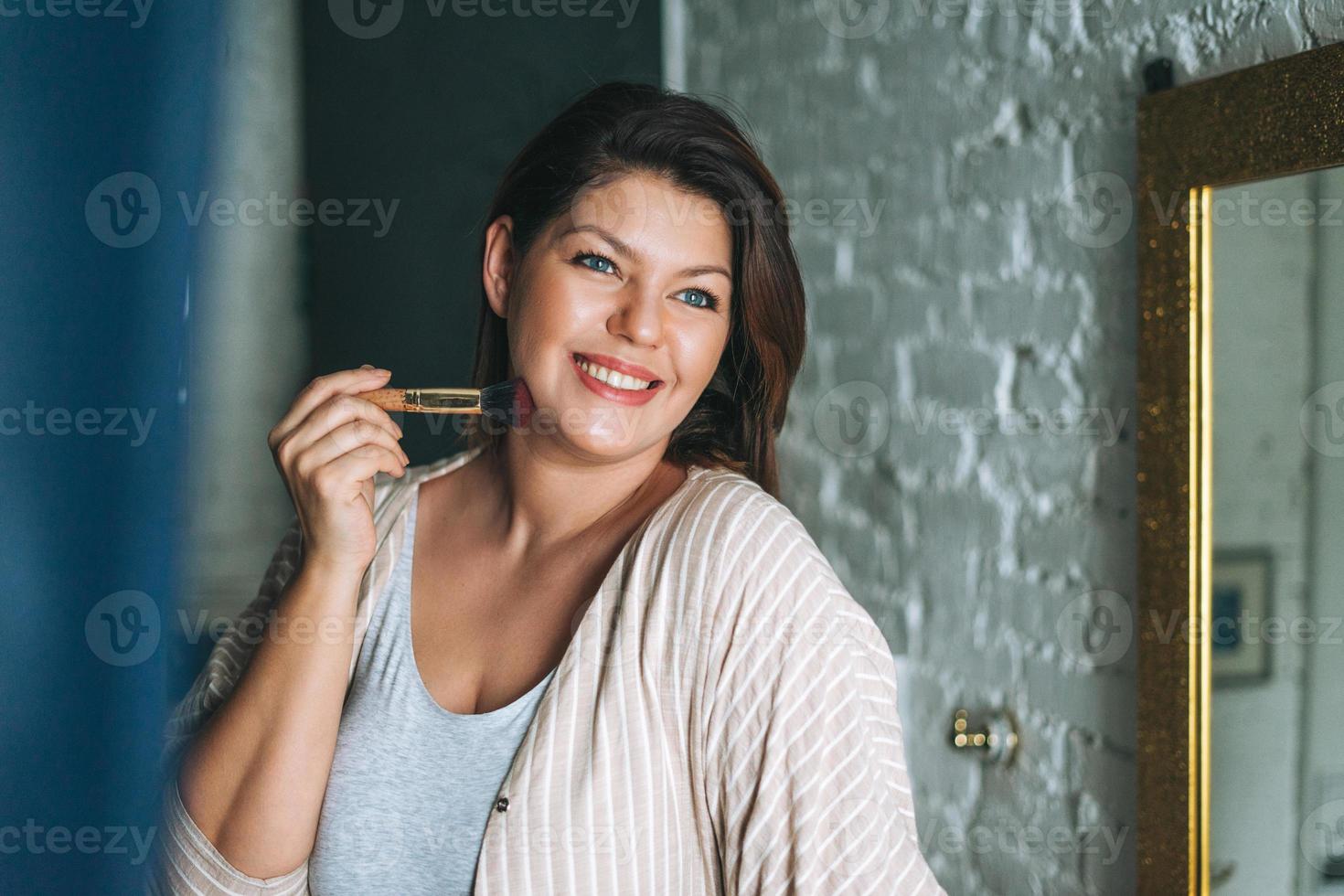 bellissimo sorridente giovane donna più dimensione corpo positivo nel casa Abiti con rendere su spazzola nel il bagno, bellezza mattina routine foto