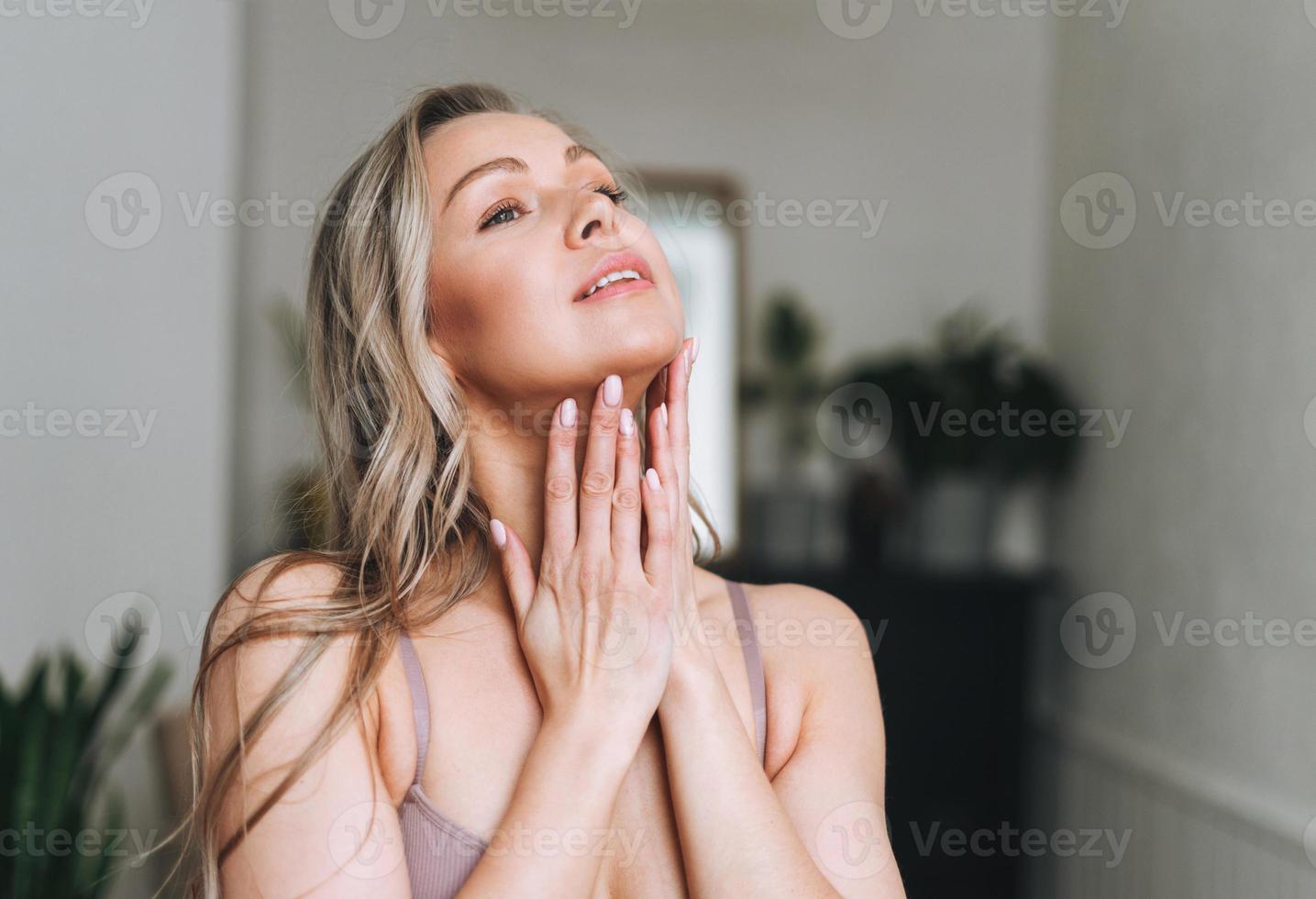 bionda sorridente donna 35 anno con pulito fresco viso e mani con lungo capelli fa facciale massaggio nel biancheria intima a bagno foto