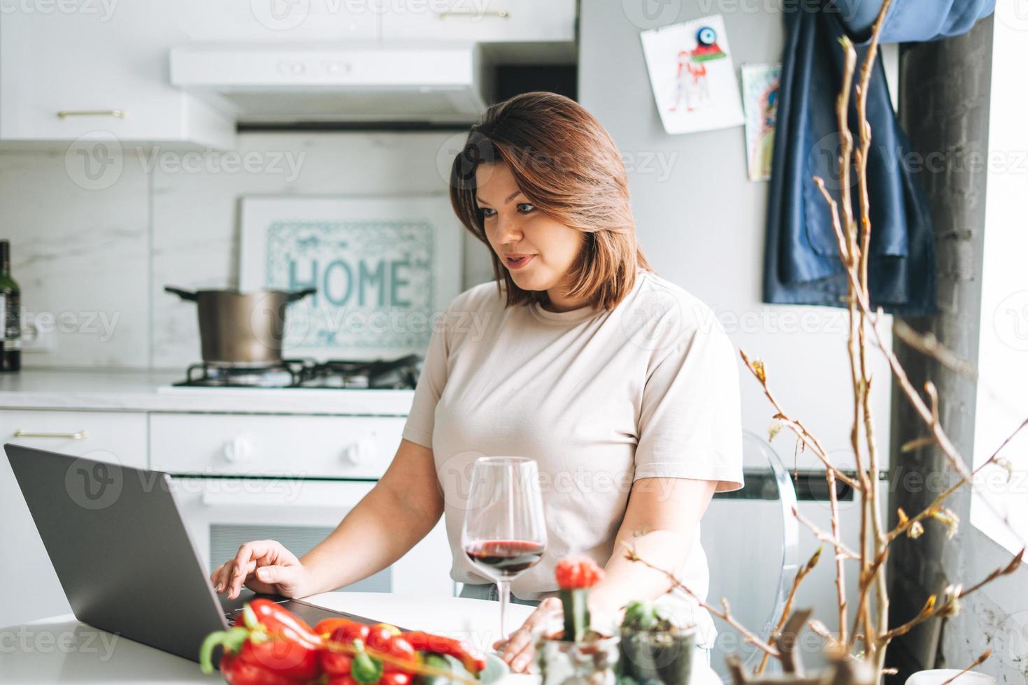 bellissimo sorridente brunetta giovane donna più dimensione corpo positivo utilizzando il computer portatile nel cucina a il casa foto