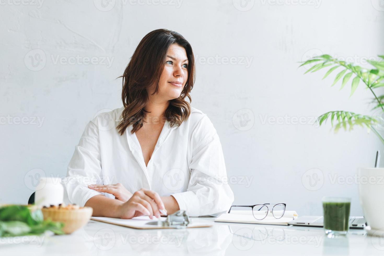 giovane sorridente brunetta donna medico dietologo più dimensione nel bianca camicia Lavorando a il computer portatile a moderno luminosa ufficio camera. il medico prescrive un' prescrizione per medicinali e vitamine a clinica foto