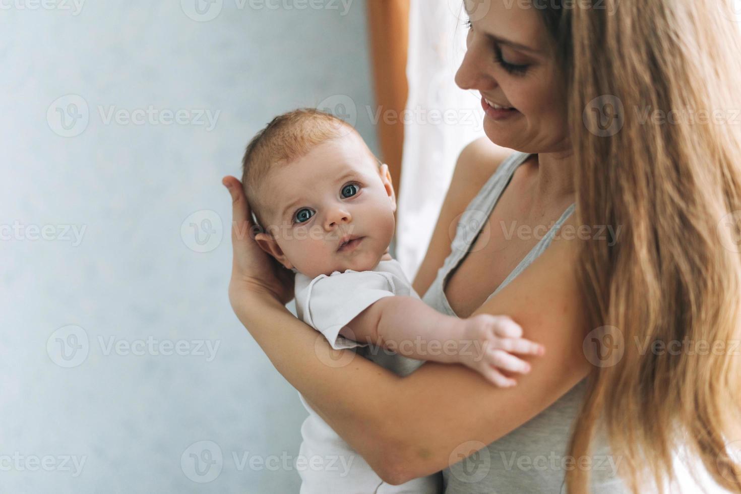 giovane donna mamma con lungo capelli Tenere bambino ragazza su mani vicino finestra a il casa foto