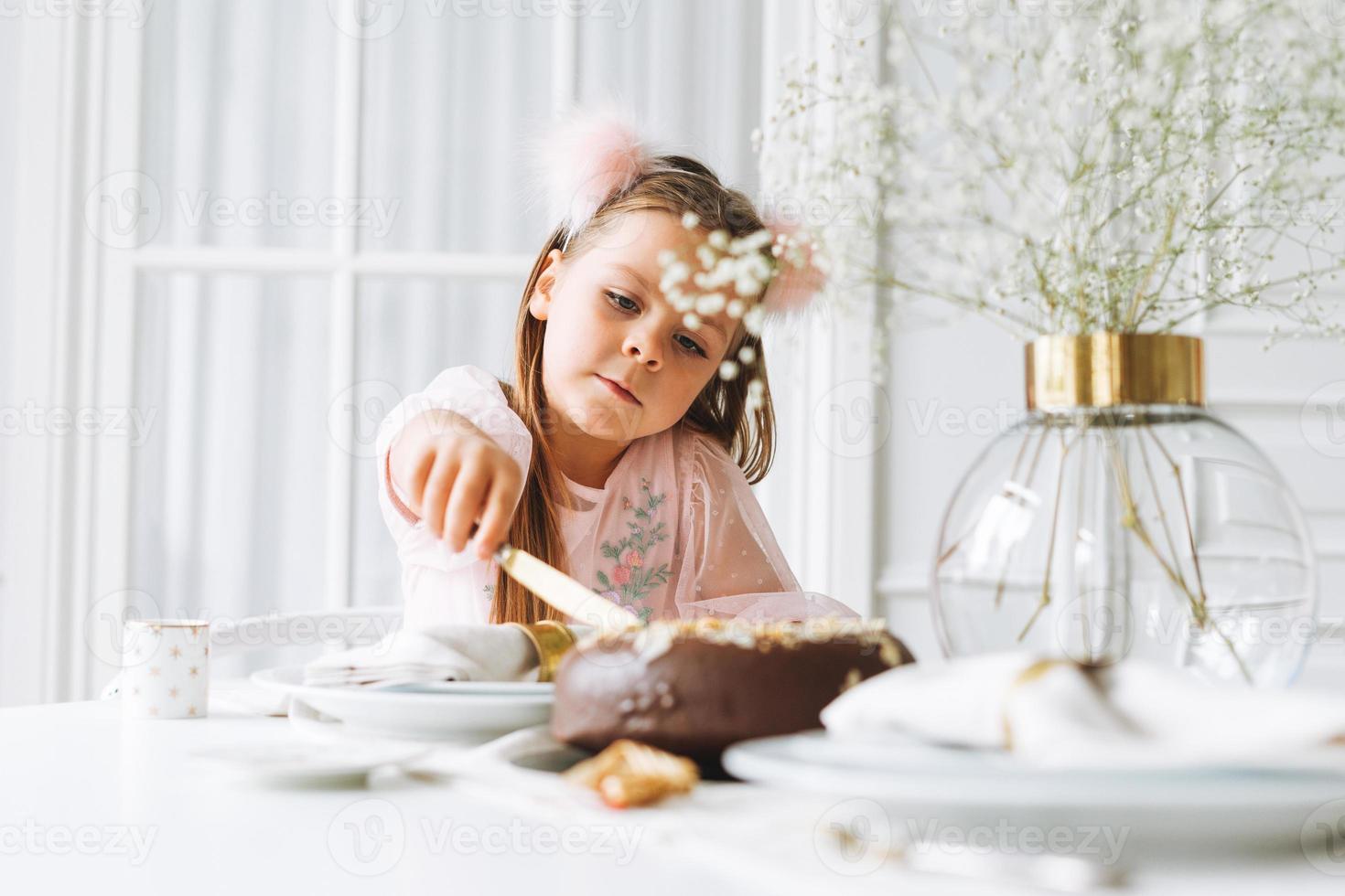 divertente carino poco ragazza con lungo capelli nel leggero rosa vestito con cioccolato torta nel mani su festivo tavolo nel luminosa vivente camera a il casa. Natale volta, compleanno ragazza foto