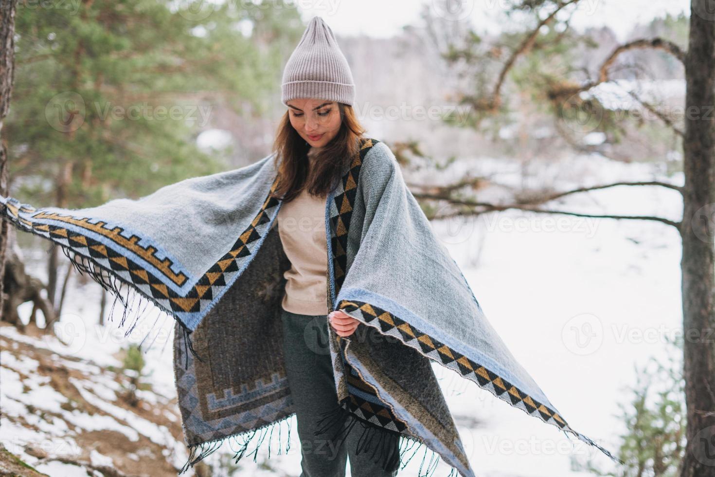 giovane brunetta bellissimo donna nel cappello e grigio poncho a piedi nel il inverno foresta foto