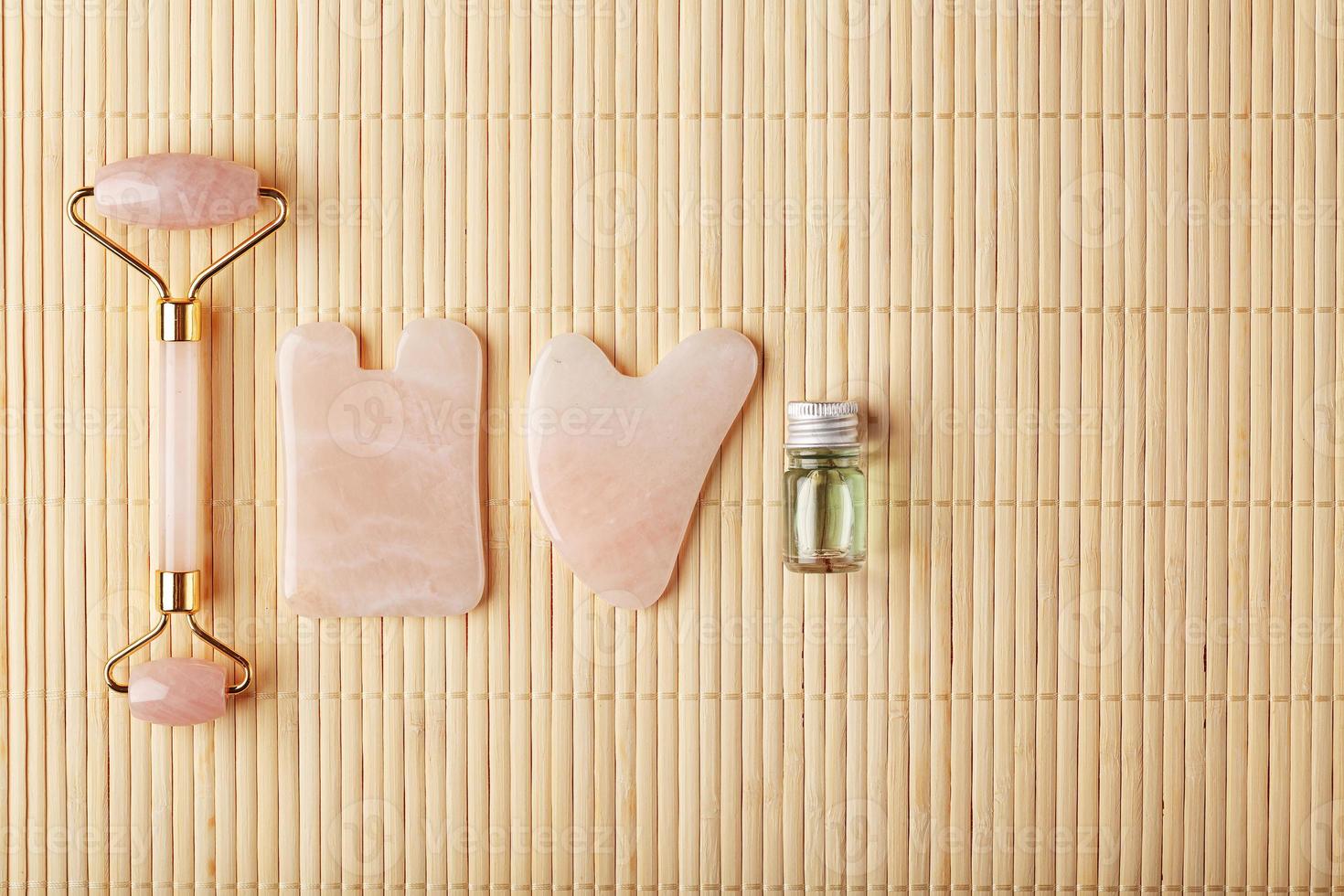 un' impostato di utensili per viso massaggio tecnica gua sha fatto di naturale rosa quarzo. rullo, giada pietra e olio nel un' bicchiere barattolo, su un' cannuccia sfondo per viso e corpo cura. foto